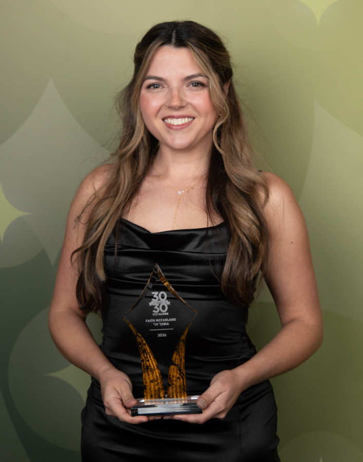 Smiling woman in a black satin dress holds a tall amber-colored glass award against a green abstract backdrop.
