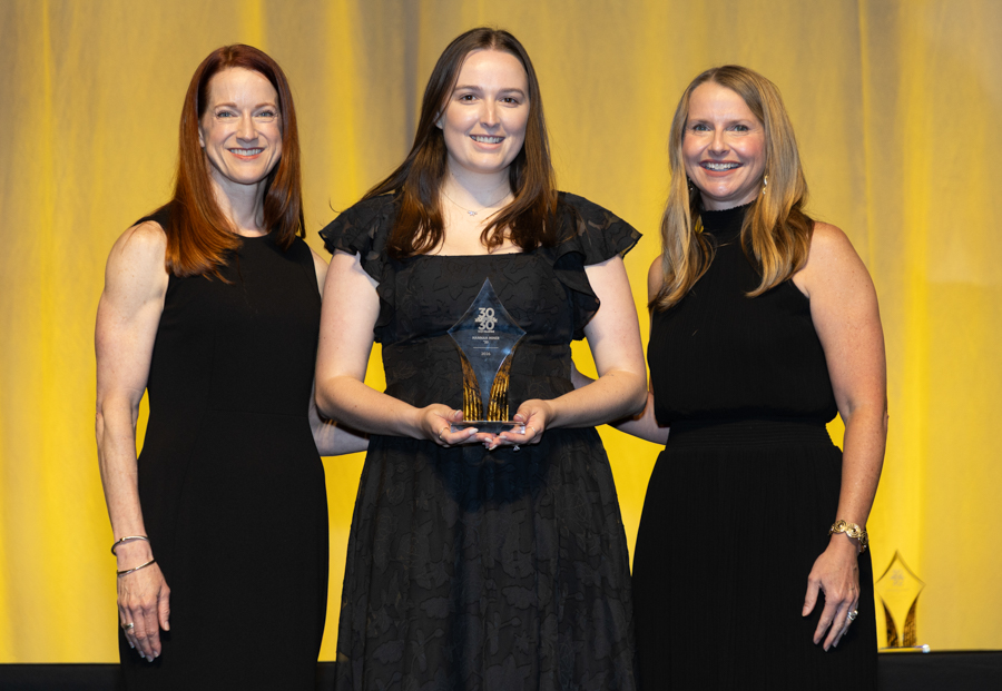 Three women in black dresses stand on a stage with a yellow backdrop; the center woman holds a crystal star-shaped award.