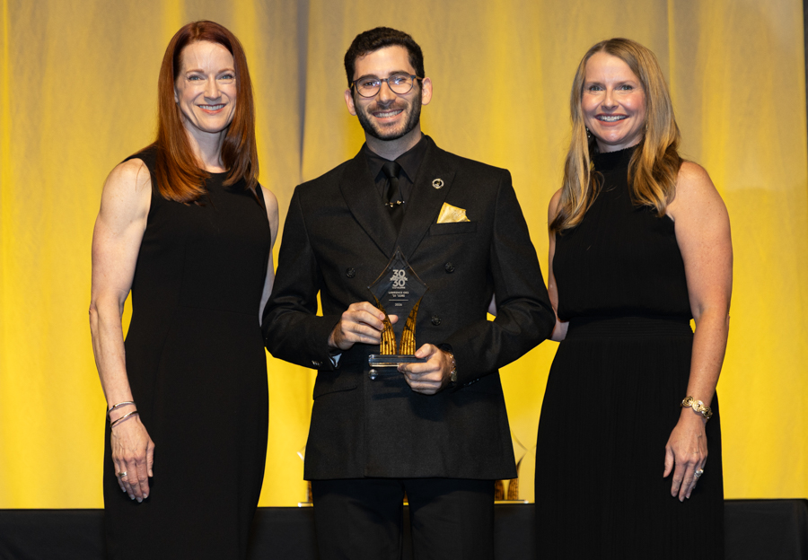 Award recipient in a black suit holding a trophy, flanked by two women in black dresses on stage under a yellow backdrop.