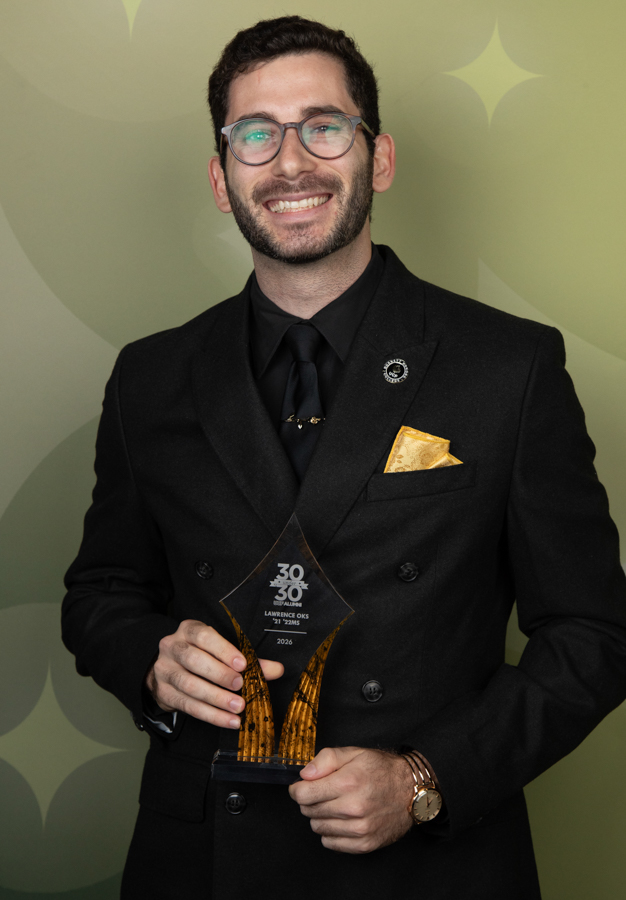 Man in a black suit smiling and holding a glass award; gold pocket square visible, starburst backdrop behind him.
