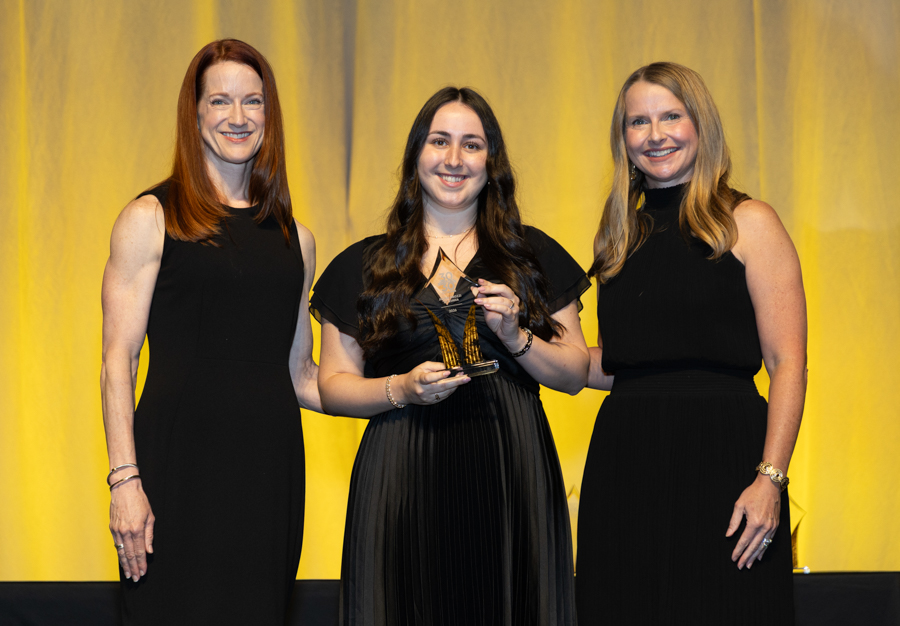 Three women in black dresses on a stage with a yellow backdrop; the center woman holds a glass award and smiles at the camera.