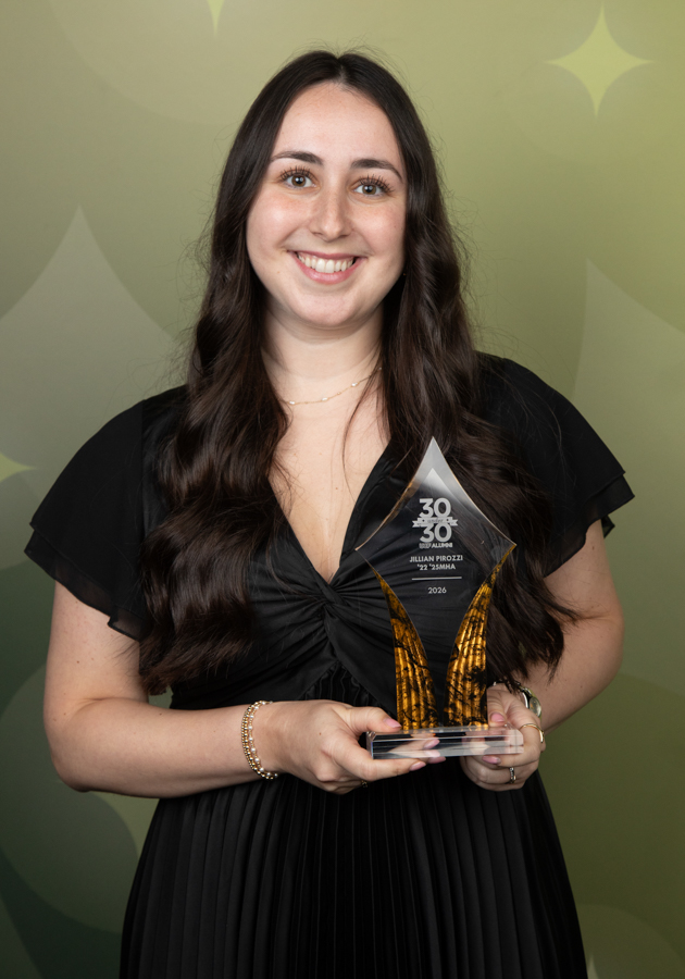 Smiling woman in a black dress holds a tall crystal trophy with gold accents, standing in front of a green patterned backdrop.