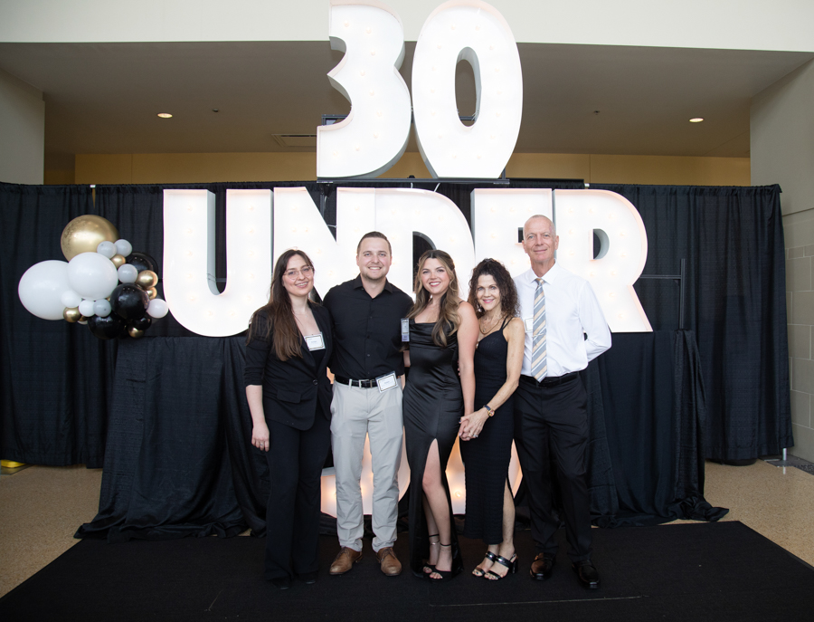 Five adults in formal attire pose together at a '30 Under' event in front of a large illuminated backdrop with black drapes and a balloon cluster on the left.
