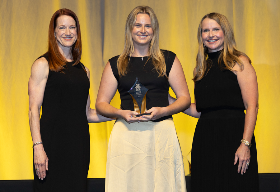 Three smiling women in black dresses on a stage; the center woman holds a glass trophy.
