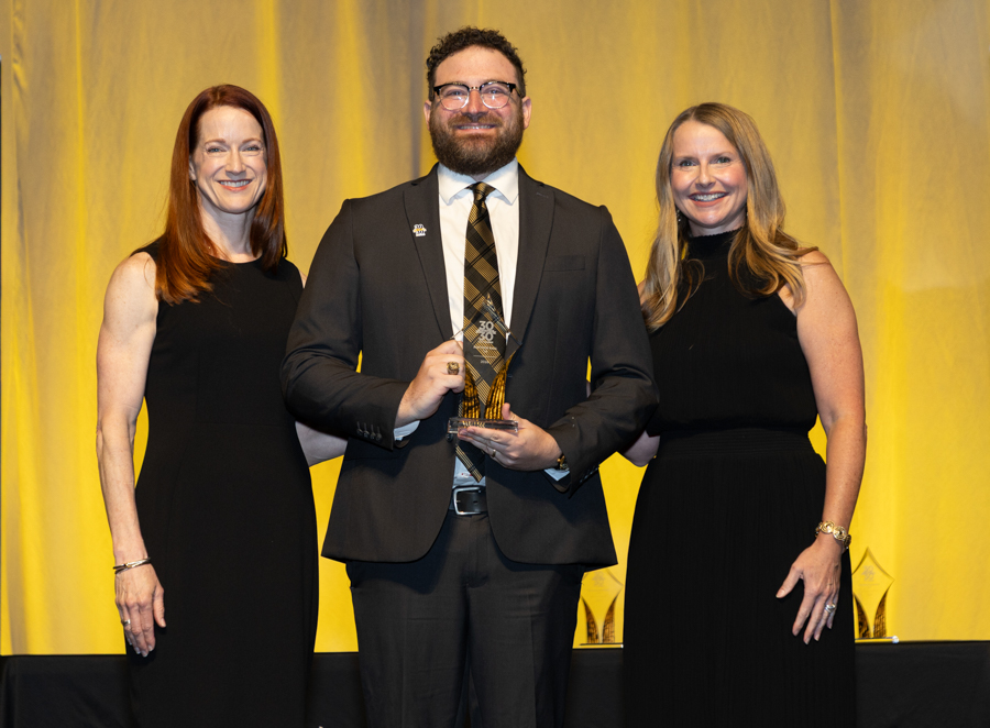 Man in a suit holding a glass trophy, flanked by two women in black dresses at an awards ceremony on a stage.