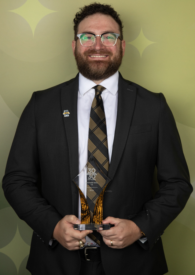 Smiling man in a black suit and plaid yellow tie holding a glass award against a green decorative backdrop.