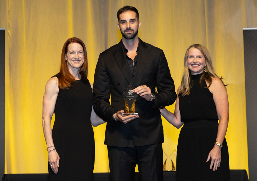 Three adults in formal black attire pose on a stage with a yellow backdrop, the man in the center holding a glass trophy with a gold base.