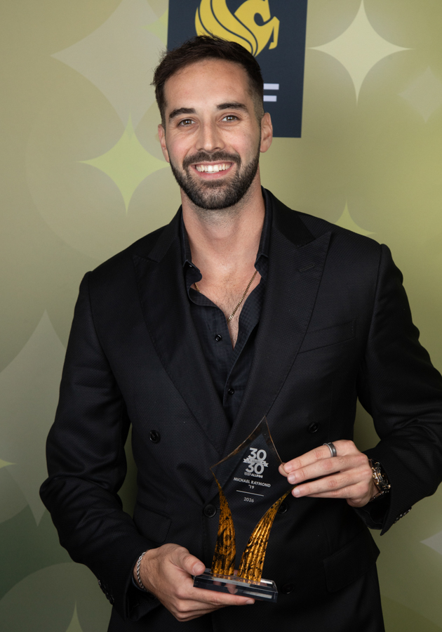 Smiling man in a black suit holding an award trophy at a formal event with a star-patterned backdrop.