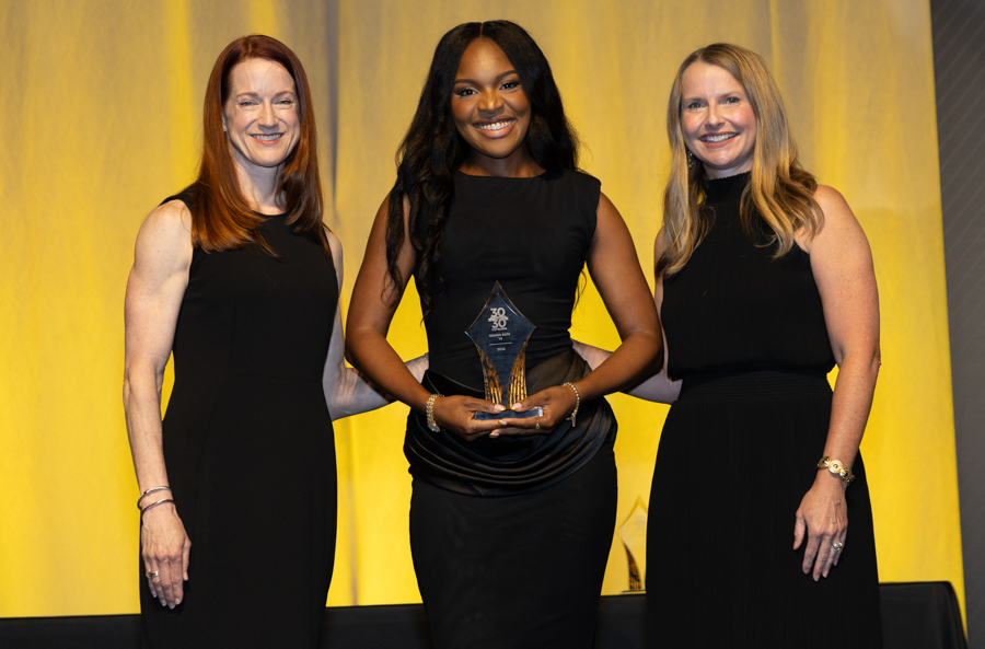 Three women in black dresses on stage, center woman holding an award trophy and smiling.