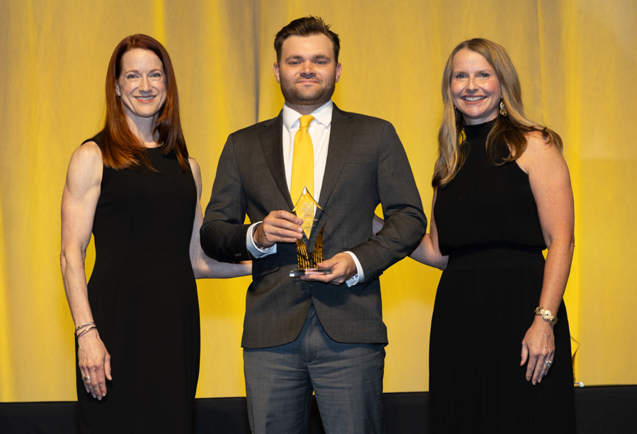 Man in a gray suit holds a glass trophy on stage, flanked by two women in black dresses against a yellow backdrop.