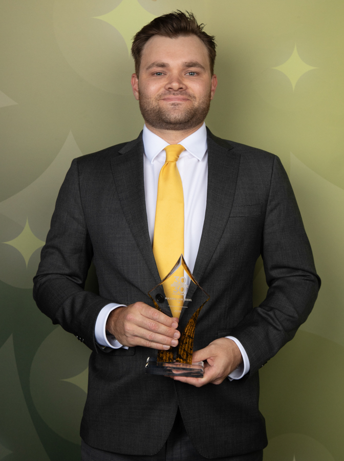 Man in a dark suit and yellow tie holds a glass pyramid trophy, smiling at the camera, against a green decorative backdrop.