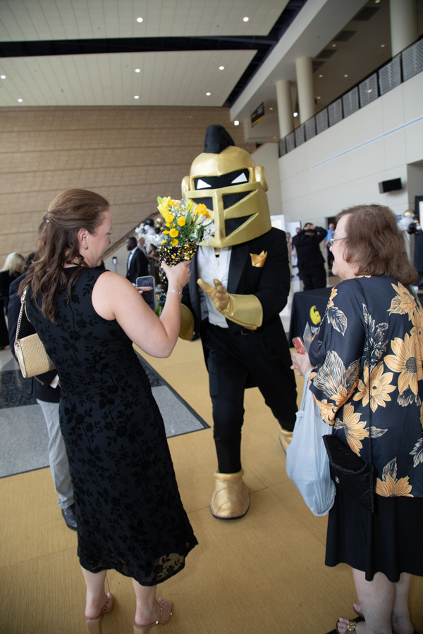 Person in a gold knight costume hands flowers to two women at a convention lobby, festive interaction