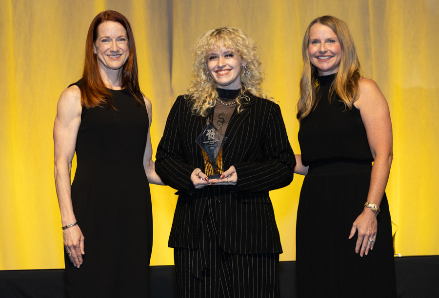 Three women in black dresses on a stage; the center woman holds a glass award against a yellow curtain backdrop.