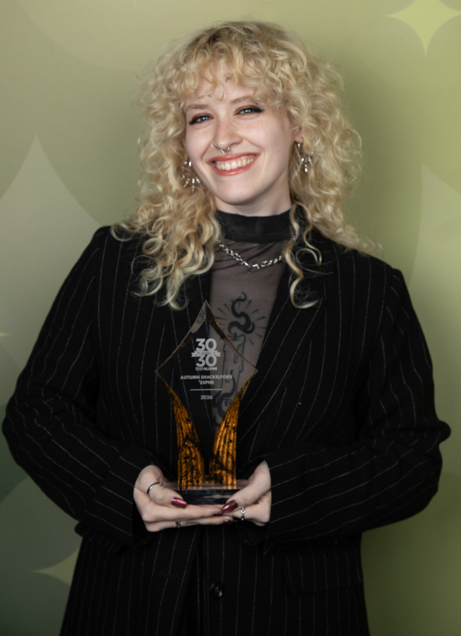 Smiling woman with curly blonde hair wearing a black pinstripe blazer, holding a glass award at a ceremony.