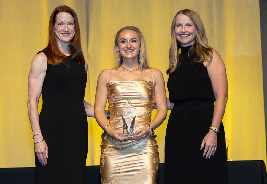 Three women in formal dresses on a stage; the center woman holds a glass award and smiles between her companions.",