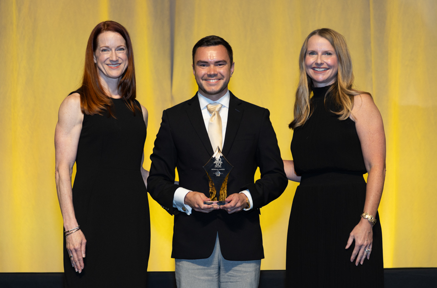 Center man in a dark suit and gold tie holding a crystal trophy, flanked by two women in black dresses on a yellow-lit stage.