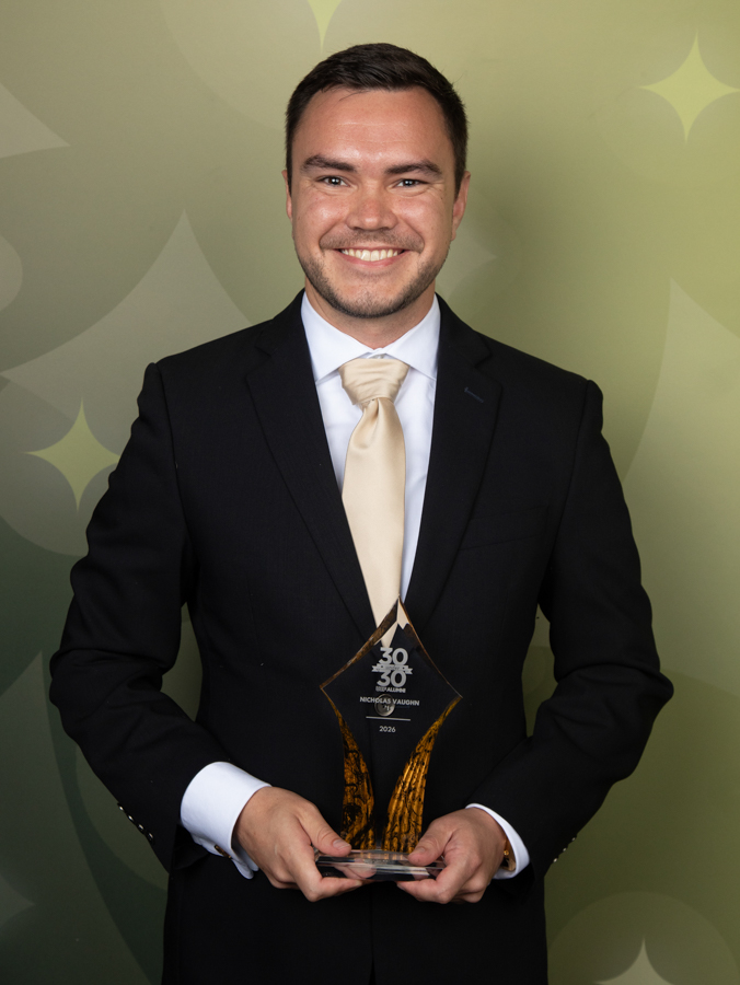 Smiling man in a black suit and beige tie holding a diamond-shaped glass trophy at an awards ceremony against a green backdrop.