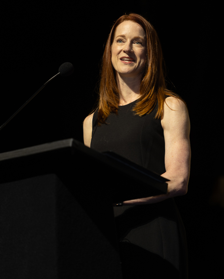 Female speaker with red hair delivering a talk at a podium with a microphone on a dark stage.