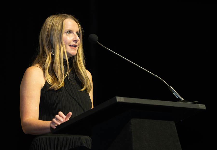 Woman in a black sleeveless dress delivering a speech at a podium on a stage with a microphone nearby