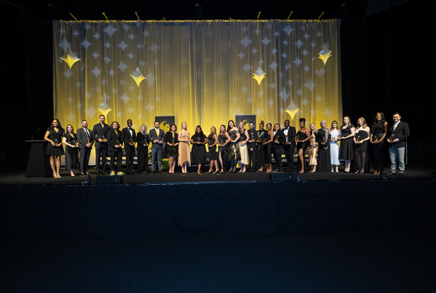 Group of men and women in formal attire on a stage, each holding awards, with a gold-draped backdrop and star decorations behind them.