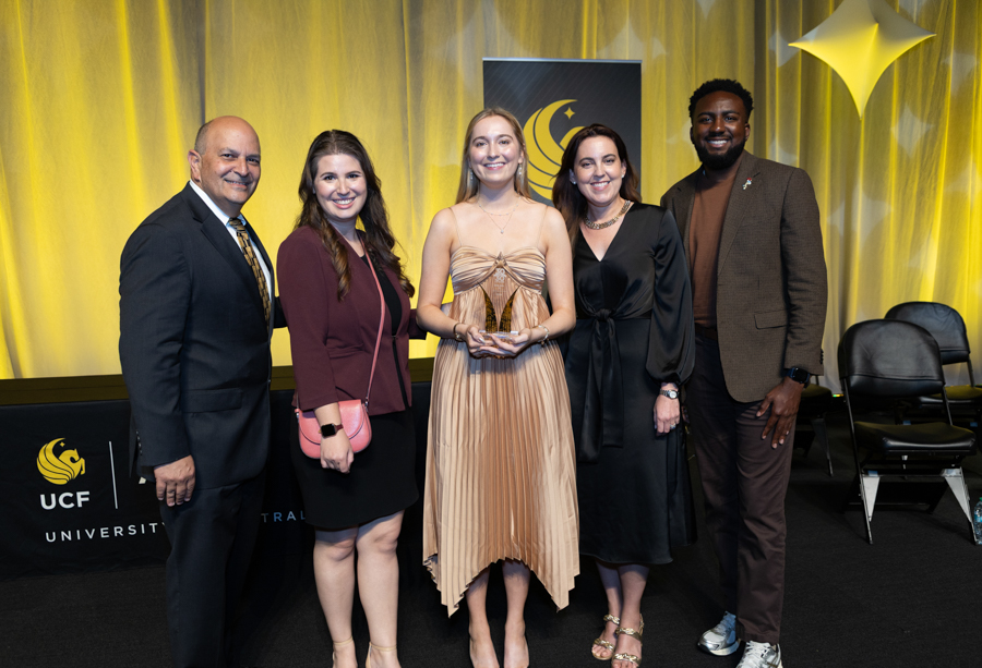 Five adults posing at a formal event; the center woman holds a trophy, with a yellow-lit stage and UCF banners behind them.
