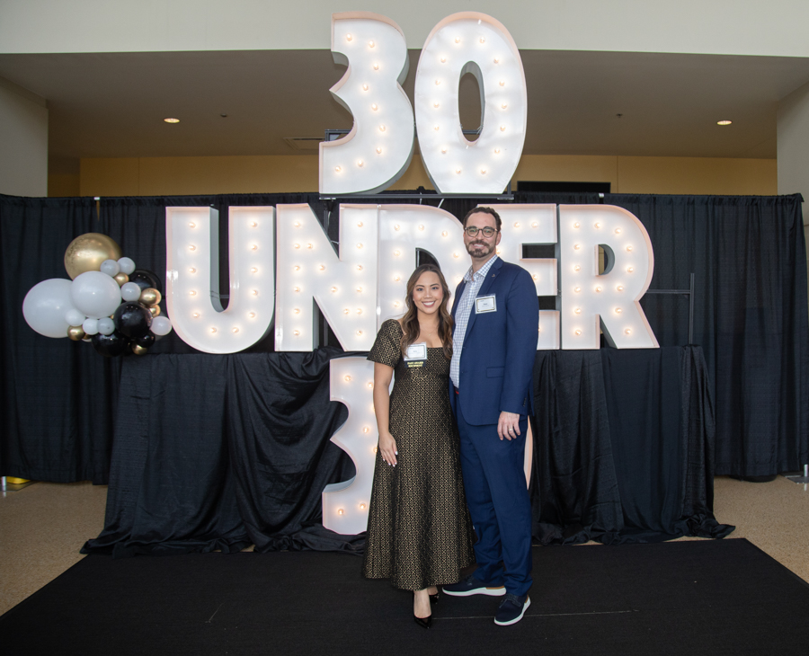 Two people pose in front of a large illuminated '30 UNDER 30' sign with balloons on the left at an awards event.