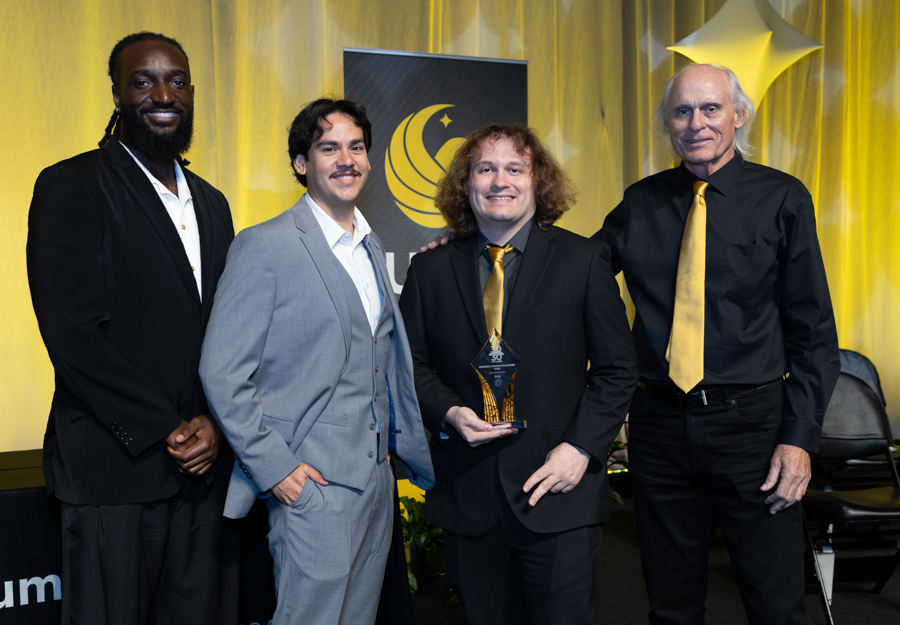 Four men in suits posing on stage at an awards ceremony; the man third from left holds a glass trophy.