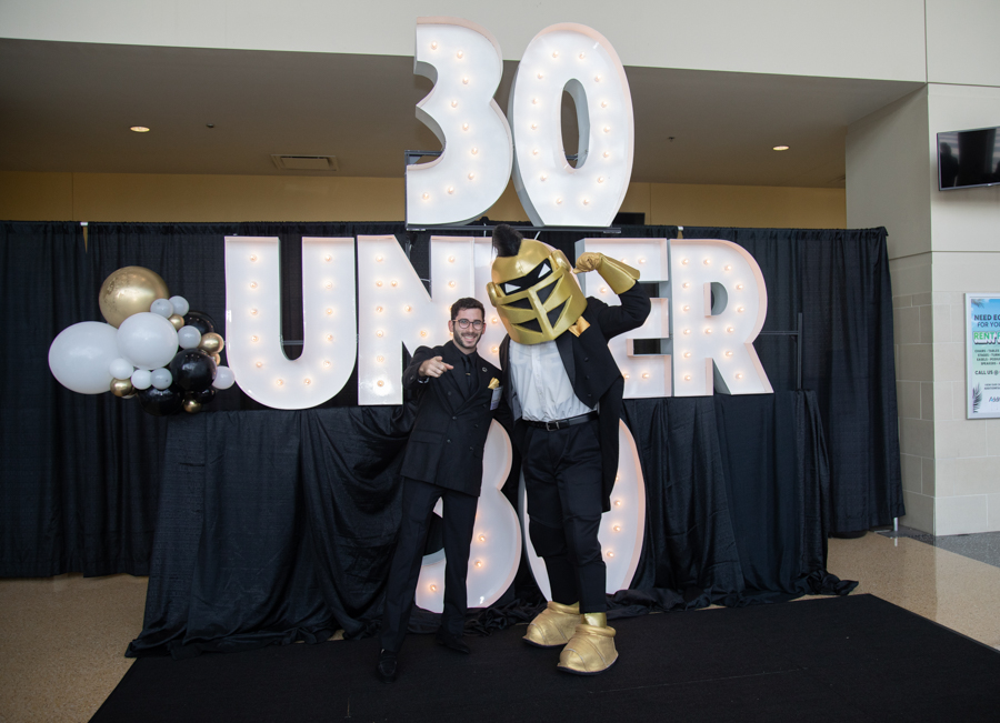 Two men posing in front of a lit '30 Under 30' backdrop; one in a black suit, the other in a gold helmet and suit, flexing their arms.