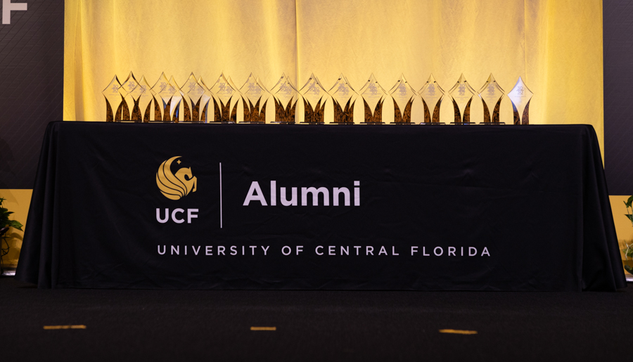 Stage table with black cloth reading 'UCF Alumni' and 'University of Central Florida' beside a row of glass trophy awards at an alumni event.