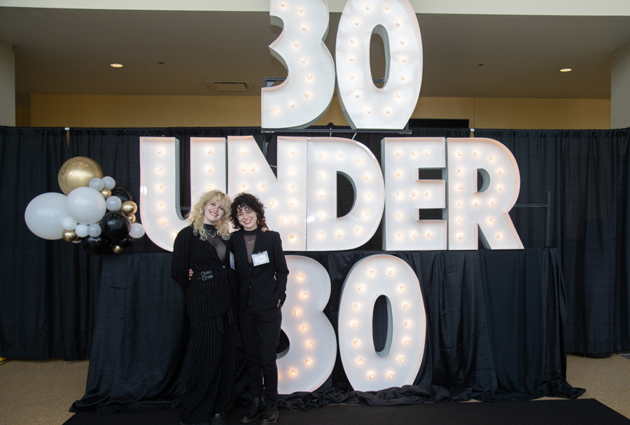 Two women pose in front of a large illuminated '30 UNDER 30' sign with a black curtain backdrop and a balloons cluster on the left.
