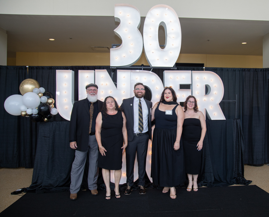 Five adults in formal attire pose in front of a large illuminated '30 UNDER' sign with balloons behind them at a event backdrop.