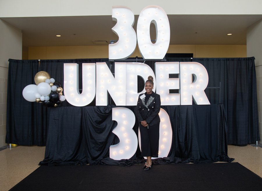 Woman in a black suit posing in front of oversized illuminated '30 UNDER 30' letters with black drapes and a cluster of balloons on the left.