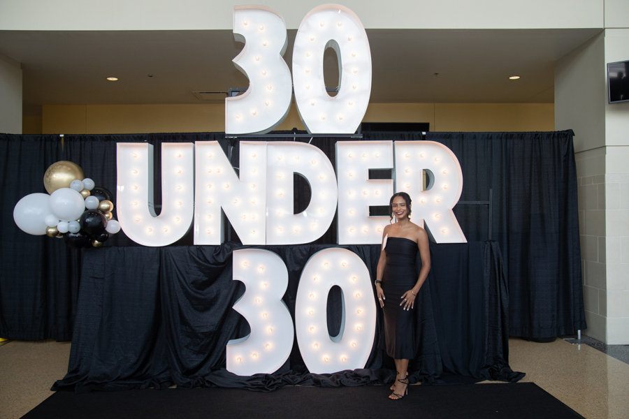Young woman in a black dress standing beside oversized illuminated '30 UNDER 30' sign with white and gold balloons on the left.