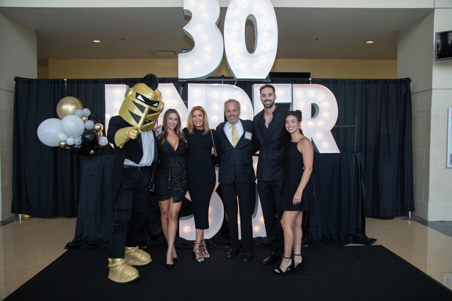 Group photo at a '30 Under 30' awards event: six people in formal attire pose on a black carpet with a costumed mascot and illuminated signage behind them.