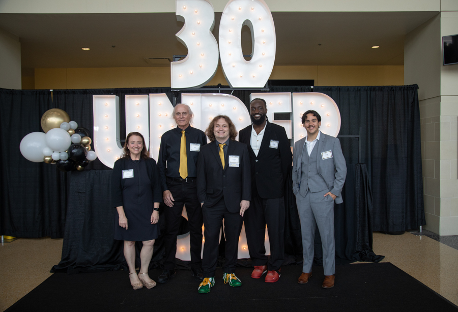 Five professionally dressed individuals pose for a photo at a '30 Under' event, with a lit sign and balloons in the background.