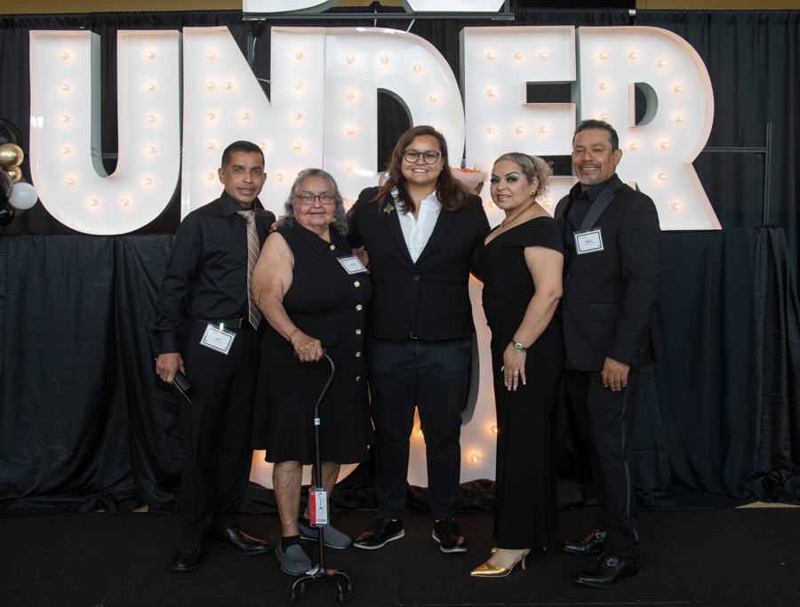 Five people in formal attire pose together in front of large illuminated letters at a gala, with one woman using a cane.