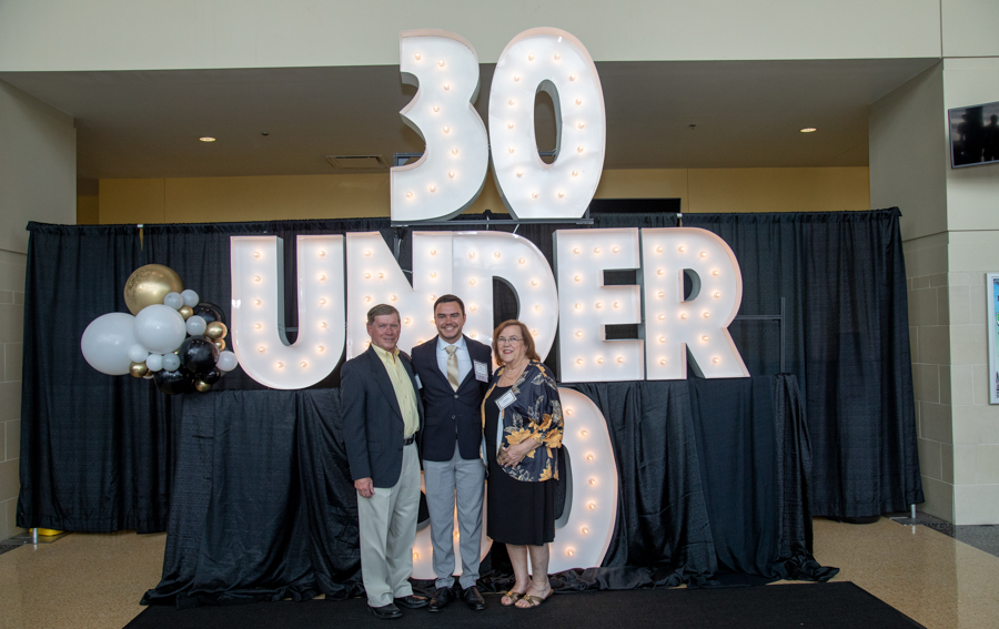 Three people pose in front of a large illuminated '30 UNDER 30' sign at a black-draped event backdrop with balloons on the left.
