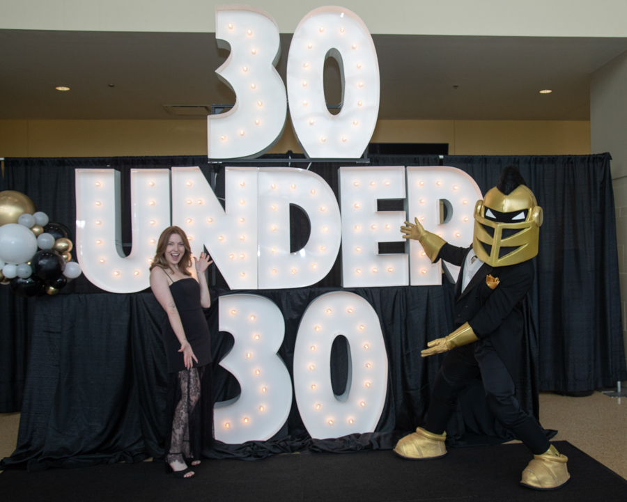 Celebration backdrop reading '30 Under 30' with a woman in a black dress posing beside a gold armored performer and balloons on the left.