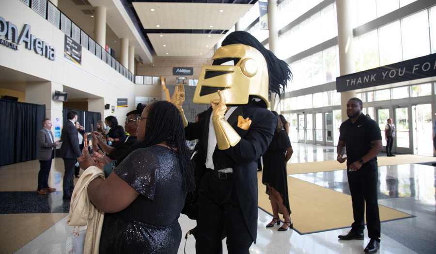 Person in a gold knight helmet mascot posing with attendees in a convention center lobby.