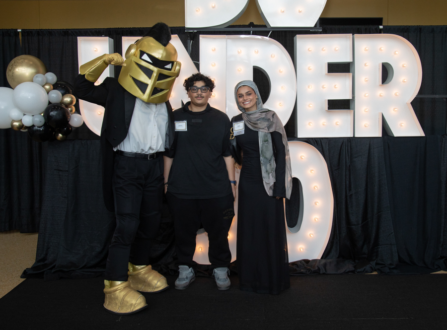 Three people pose for a photo at an event in front of large illuminated letters; left person wears a gold knight helmet and suit, middle person in black, right person in a hijab and black dress.