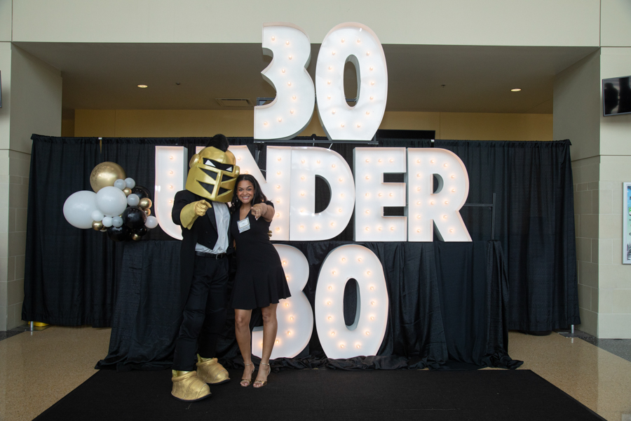 Woman in a black dress posing with a gold-armored mascot in front of a large illuminated '30 UNDER 30' backdrop and balloons at an award event.