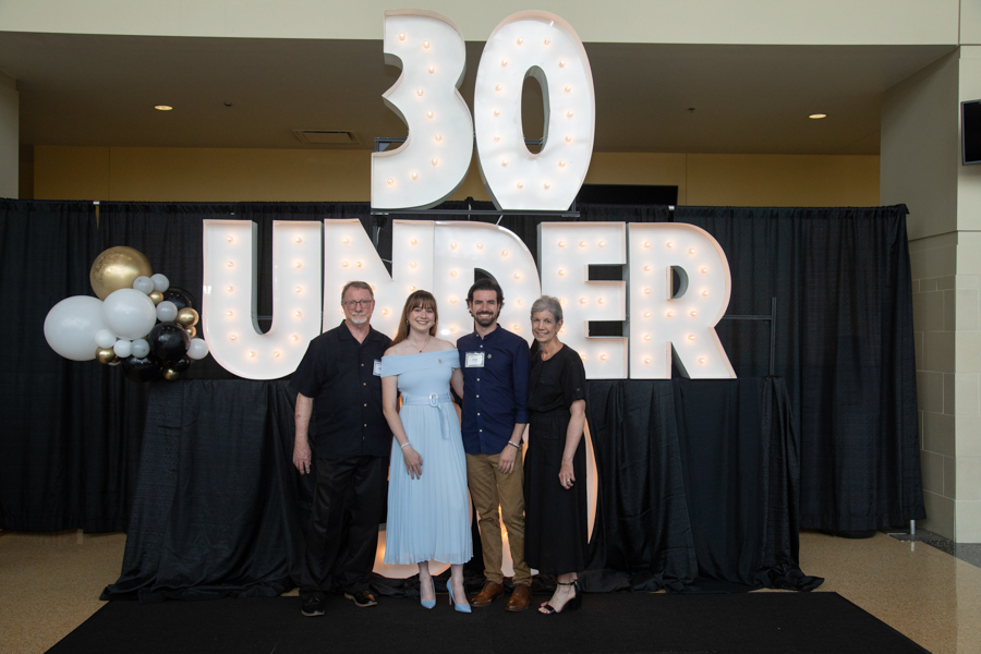 Five adults pose in front of a large illuminated sign that reads '30 UNDER' at an indoor event, with balloons on the left and a black backdrop.