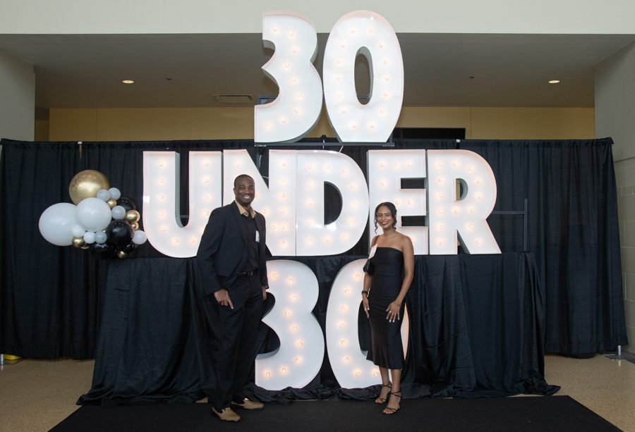Two people pose in front of a large illuminated '30 UNDER 30' display at an event with black drapes and balloon decor.