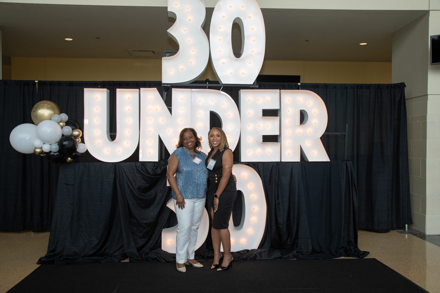 Two women pose in front of a large illuminated '30 UNDER 30' sign at a celebration with black drapes and balloons nearby.