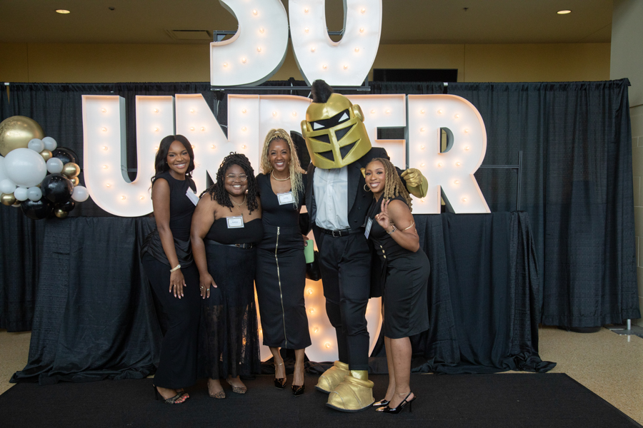 Five women in black dresses pose with a gold knight mascot in front of lit, oversized letters behind.