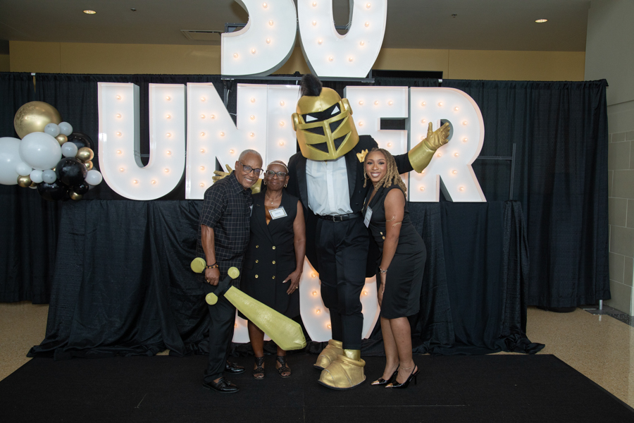 Group of four posing with a person in a gold knight costume under illuminated '30 UNDER' letters.