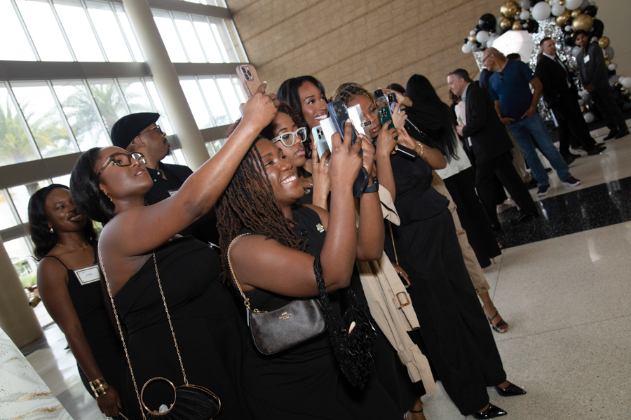 Group of people in black attire taking selfies with smartphones at a gala, balloons visible in the background.