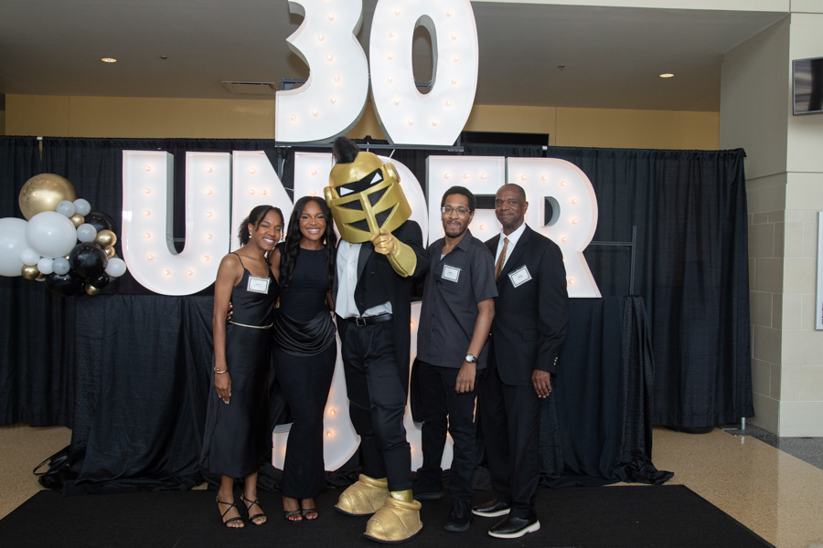 Five adults pose with a person in a gold knight costume at a 30th anniversary event, in front of a large illuminated “30” sign and black backdrop with balloons.