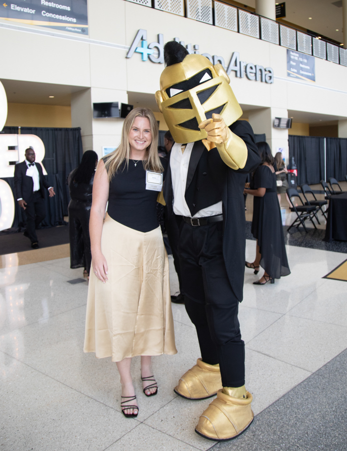 Smiling woman in a black top and beige skirt poses with a gold knight mascot in an arena lobby.