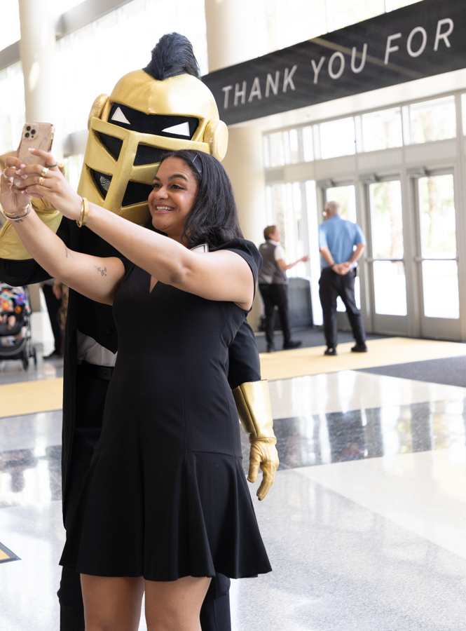Woman in a black dress takes a selfie with a person in a gold knight costume in a bright lobby setting, banner reading 'THANK YOU FOR' in the background.